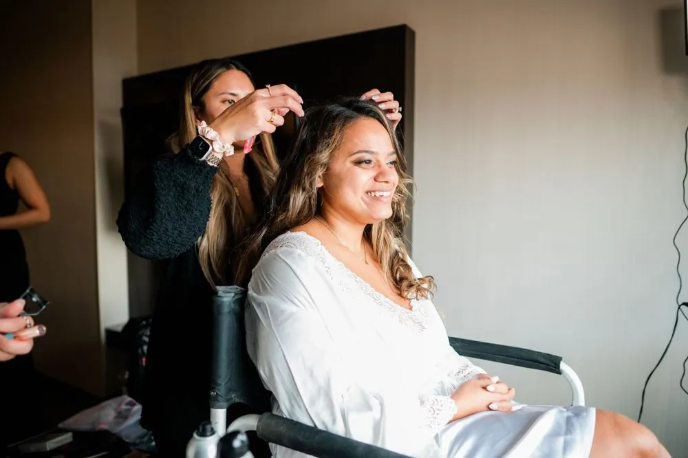 The bride smiles in a white robe as a stylist works on her hair
