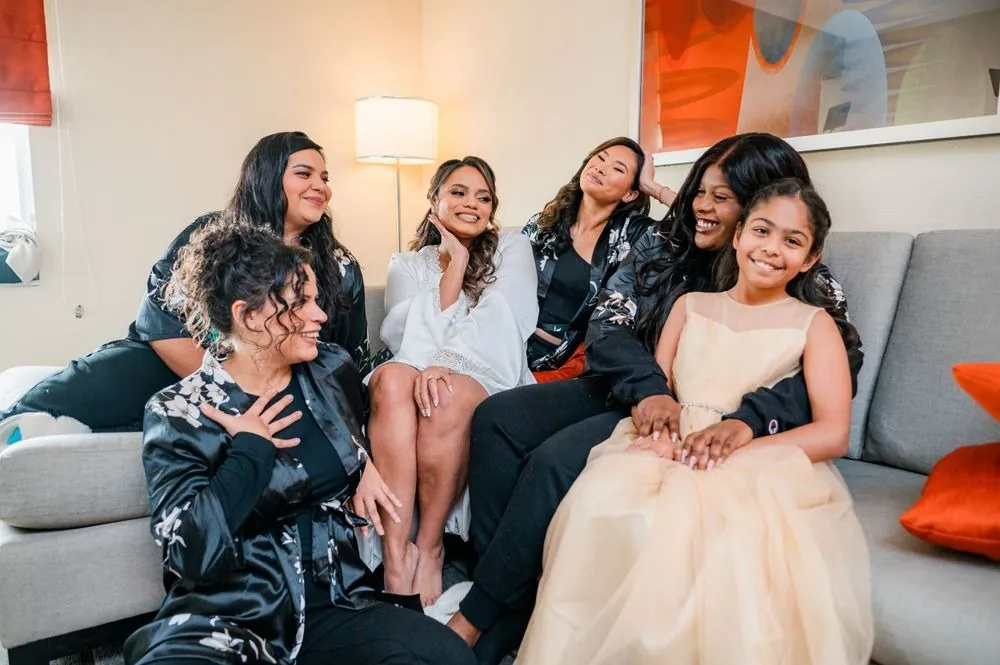 Bridesmaids and a flower girl gather on a gray sofa laughing with the bride