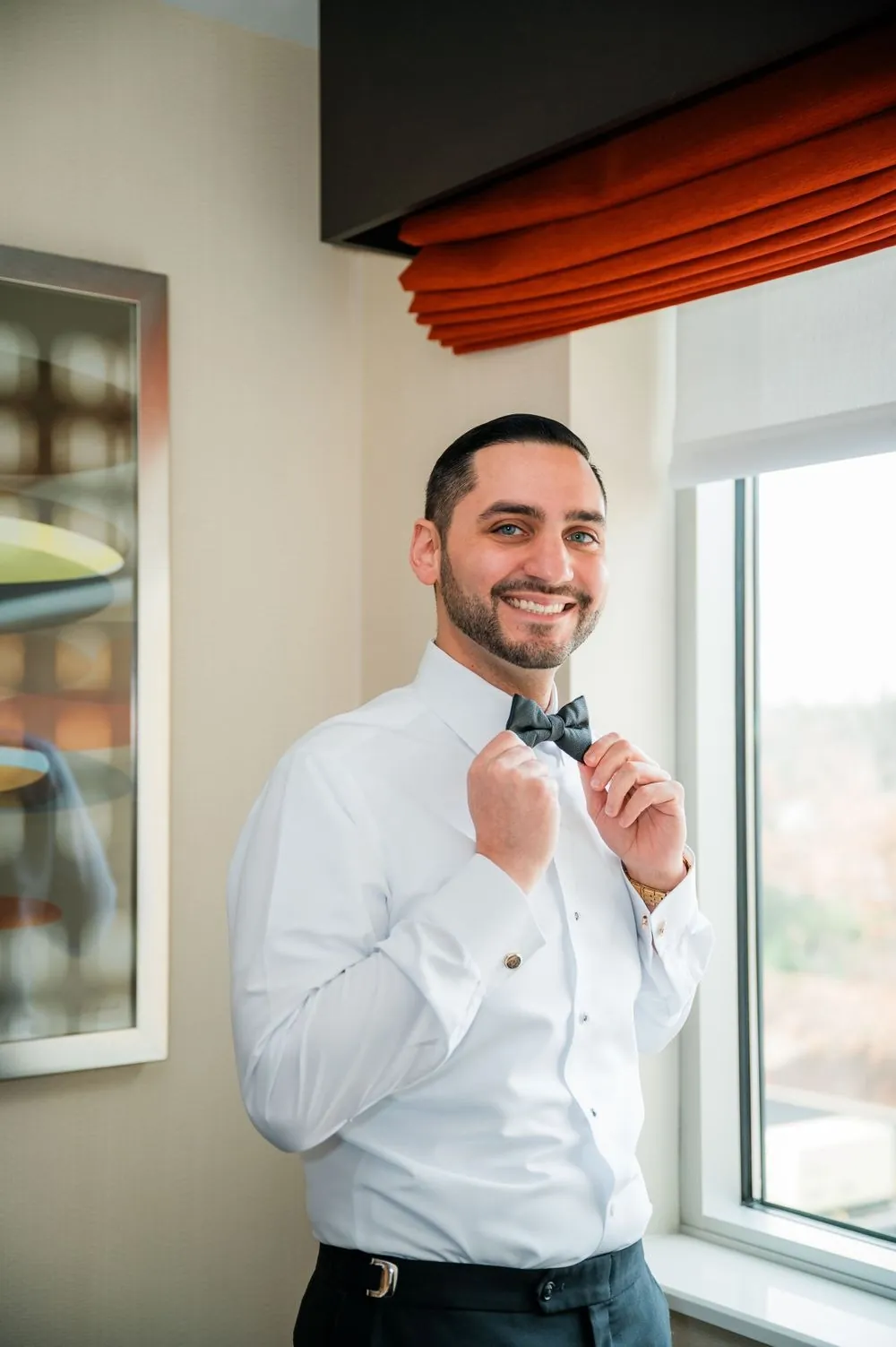 The groom adjusts his bow tie beside a bright window