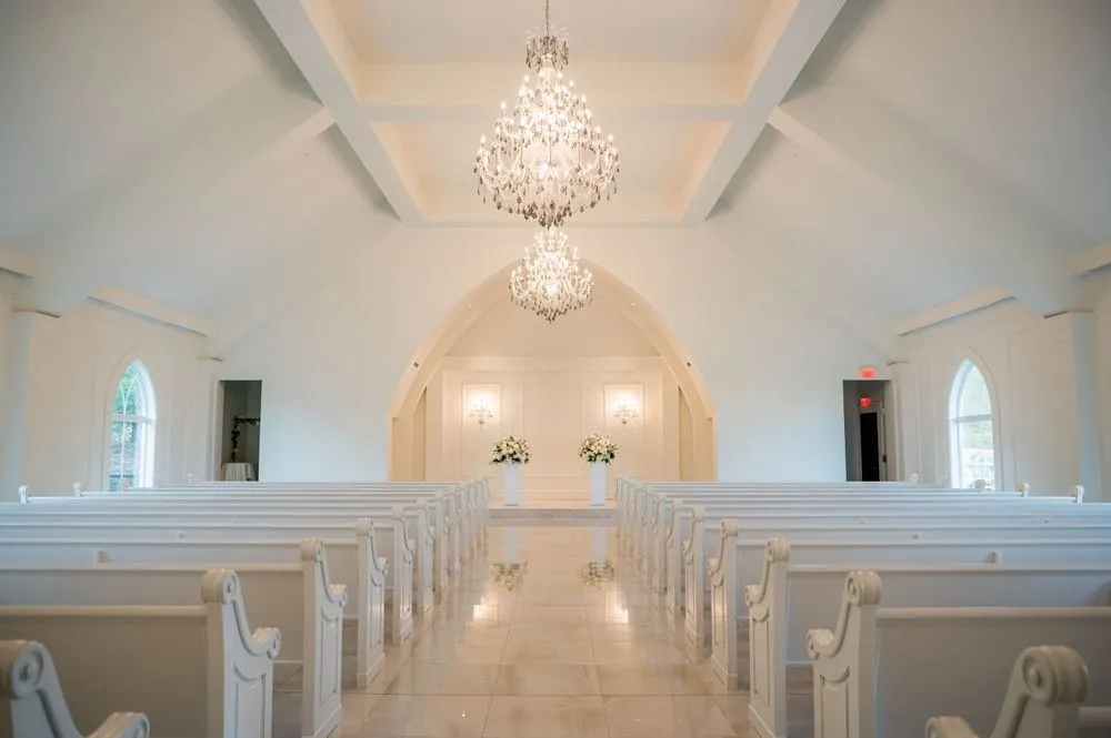 The empty white chapel with arched ceiling, chandelier and rows of white pews
