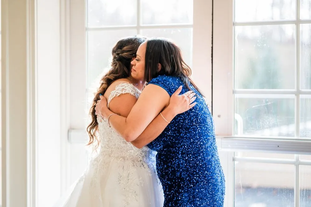 The bride hugs a woman in a sparkling blue dress near the chapel windows
