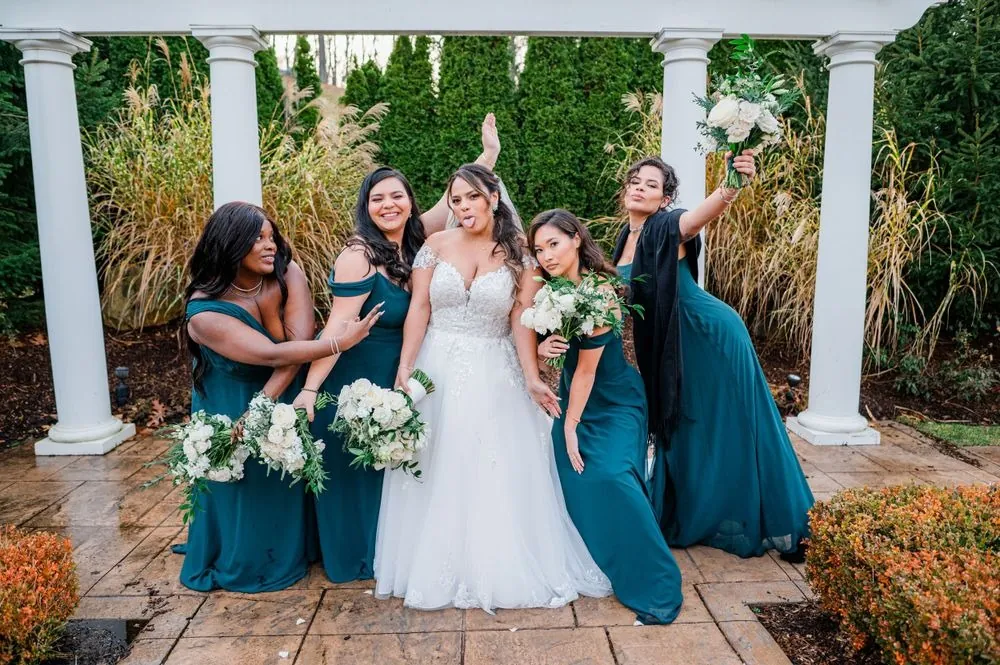 The bride laughs with bridesmaids in teal gowns beside white columns outdoors