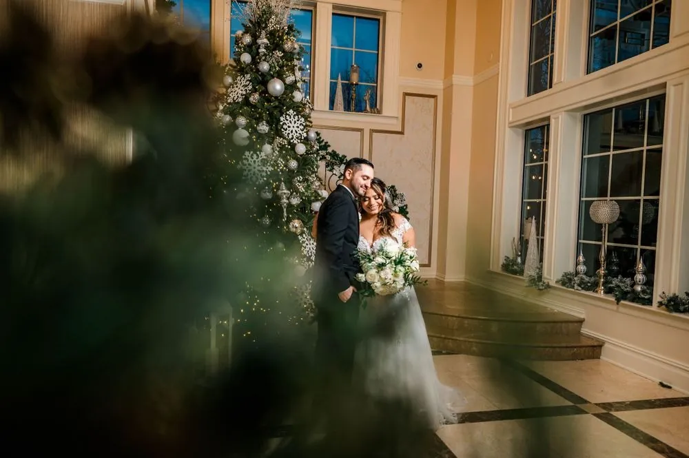 Ashley and Michael stand close beside a silver-decorated Christmas tree by tall windows