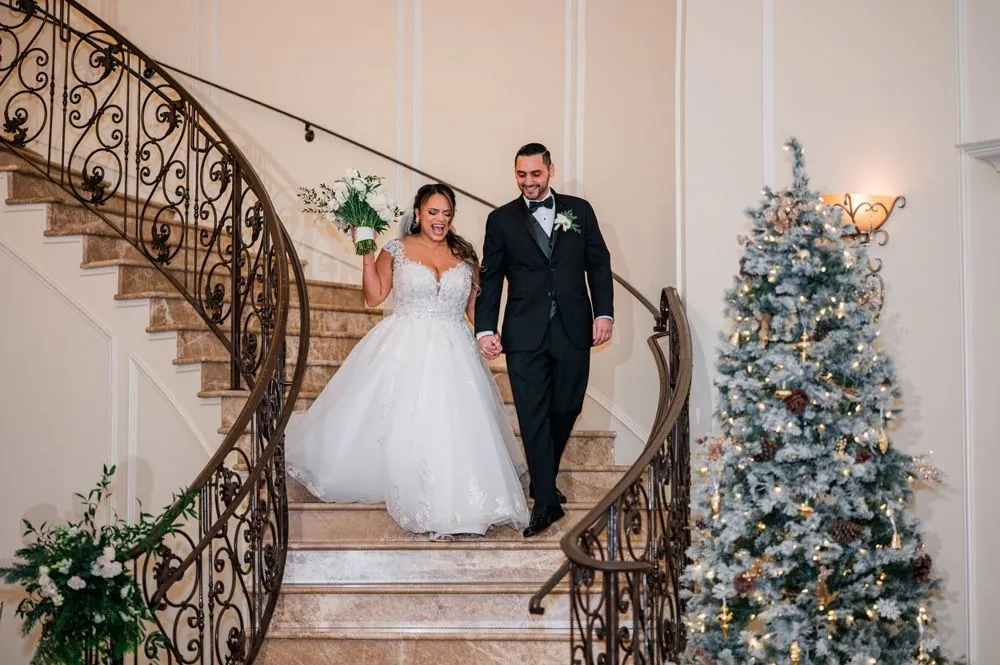 The couple enters down a curved iron staircase beside a white Christmas tree