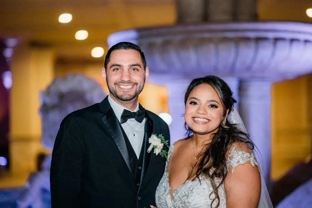Ashley and Michael smile together in front of a stone fountain under purple light