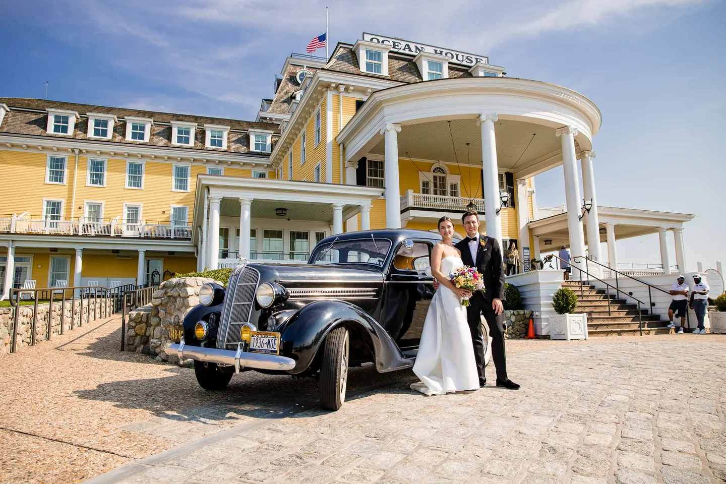 Avery and Dan stand beside a vintage black car in front of the yellow Ocean House facade