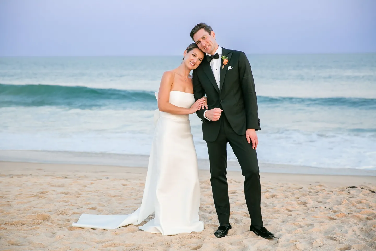 Avery rests her head on Dan's shoulder on the sand, a teal wave curling behind them