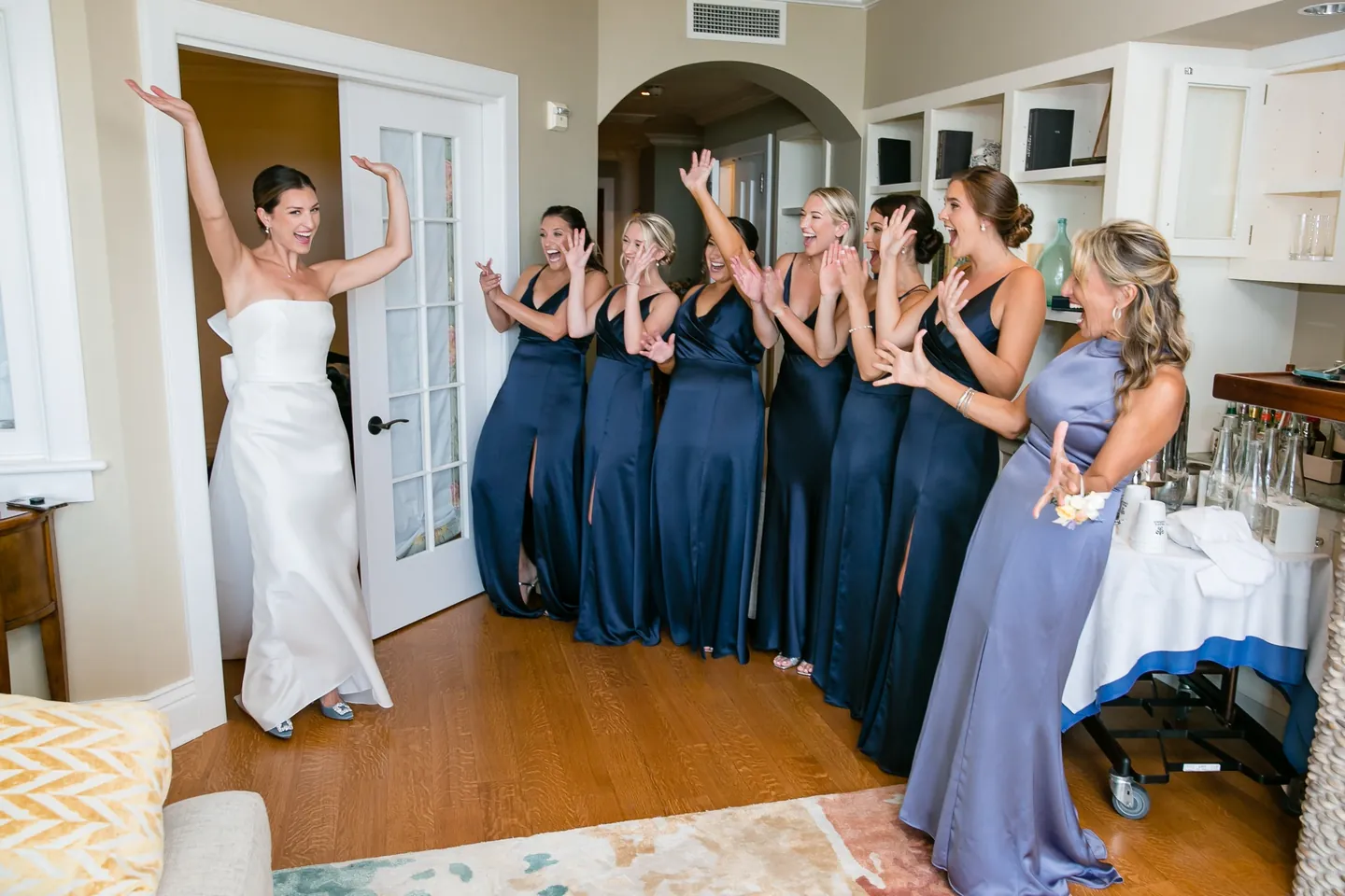 Bridesmaids in navy gowns cheer and cover their mouths as the bride walks into the room