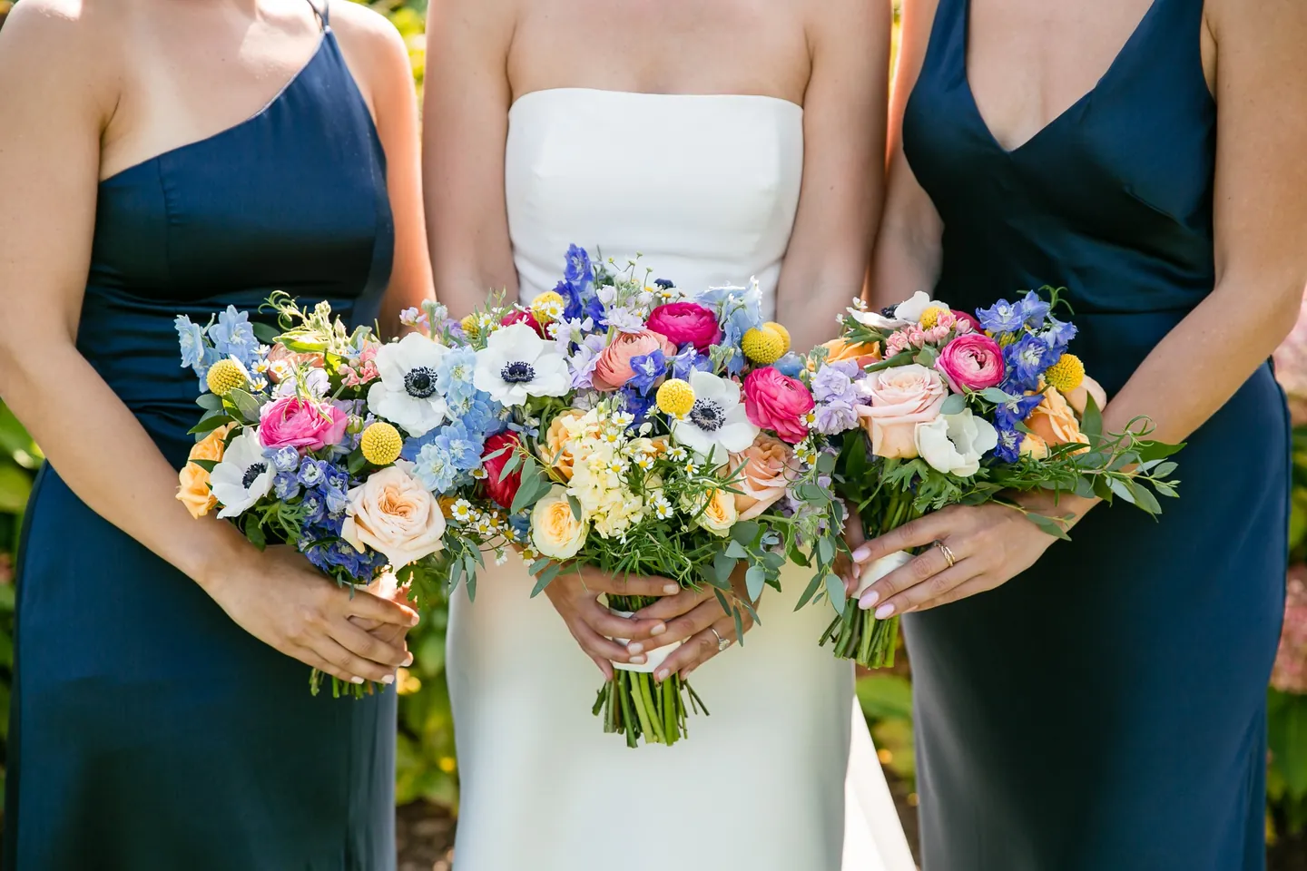 Close crop of three bouquets of anemones, ranunculus and blue delphinium held against navy dresses