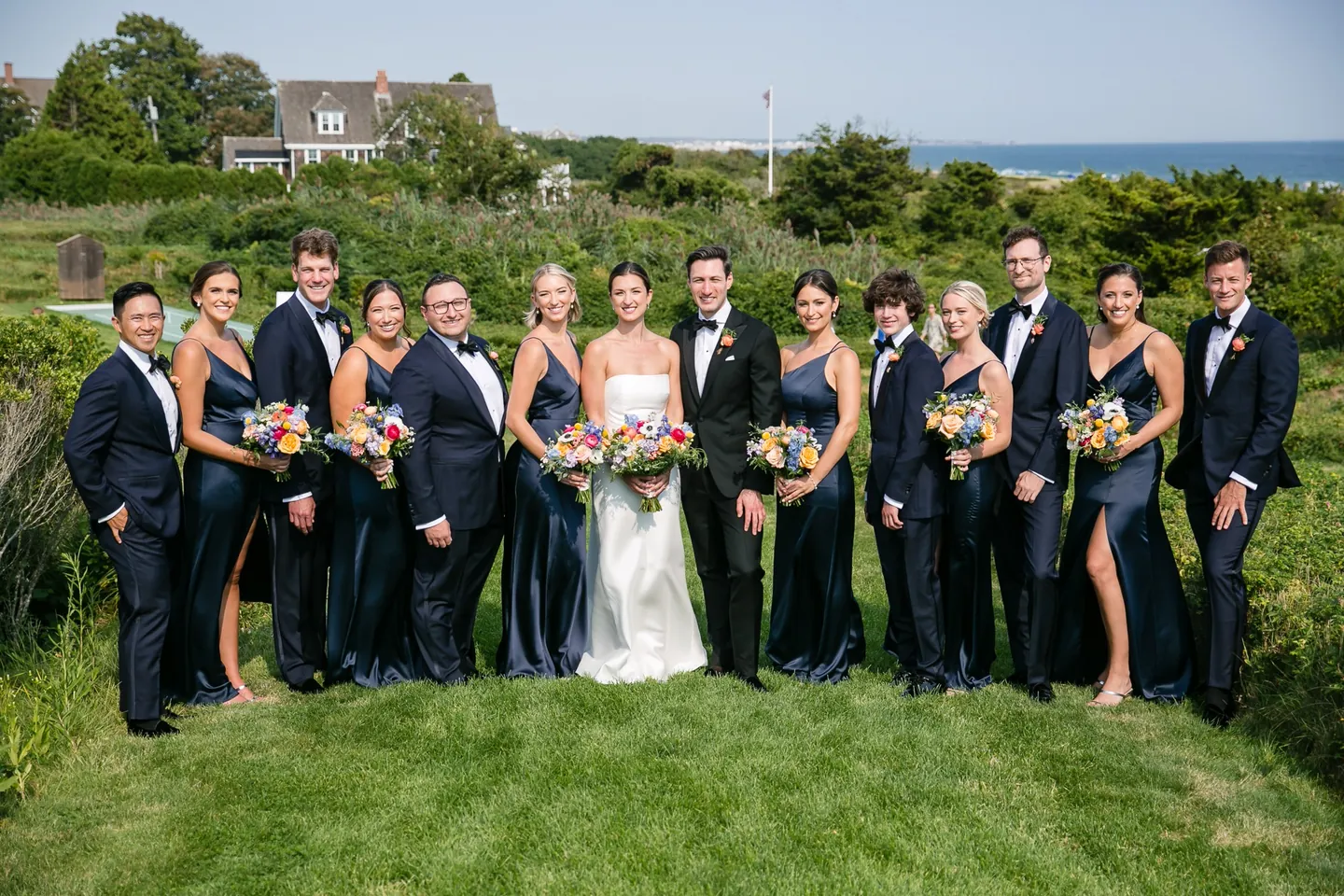 The full wedding party in navy and black tie lines up on a green lawn above the sea