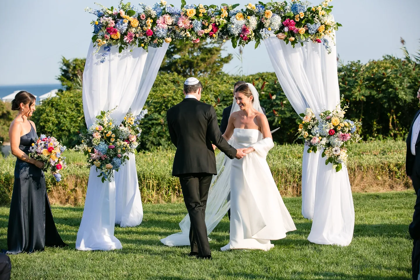 The couple stand under a white-draped arch topped with bright flowers, bridesmaids in navy to one side
