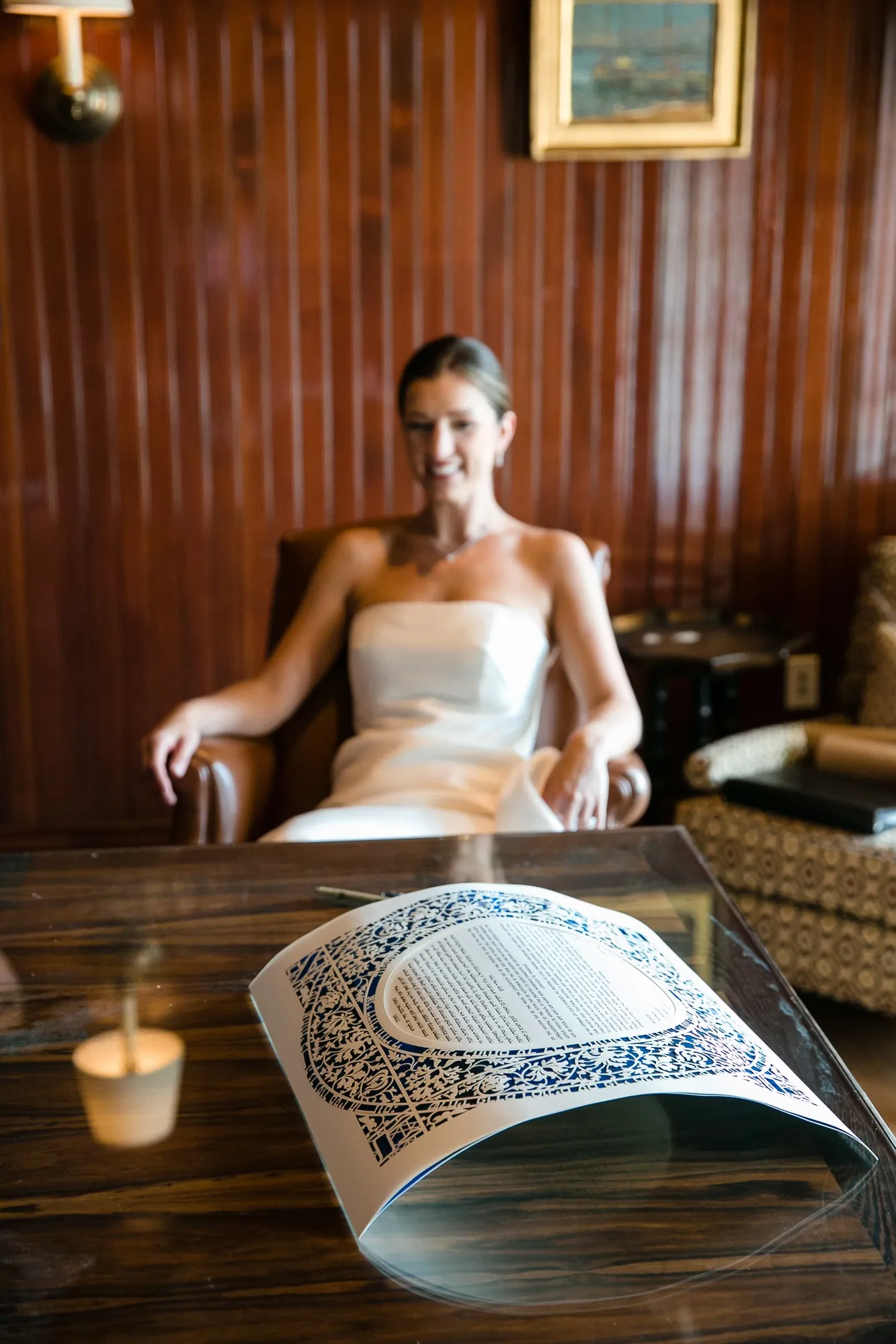 The bride sits at a wood-paneled table beside a candle with a document laid out before her
