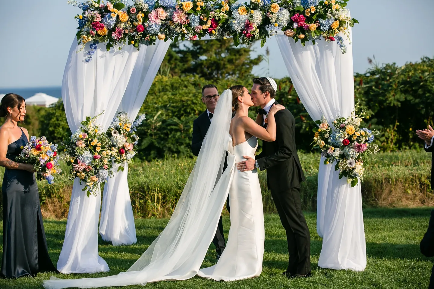 Avery and Dan kiss beneath the floral arch while the wedding party claps on the lawn