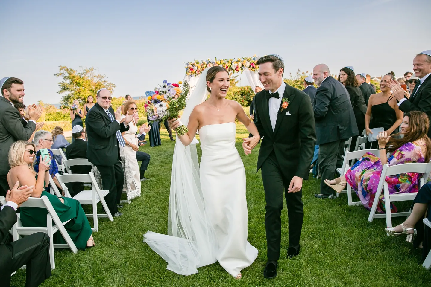 Avery and Dan walk back up the grass aisle past rows of white chairs and smiling guests