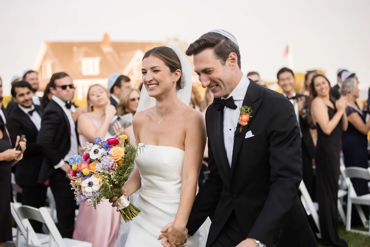 Avery and Dan laugh during the recessional, her bouquet swinging, guests in kippot applauding behind