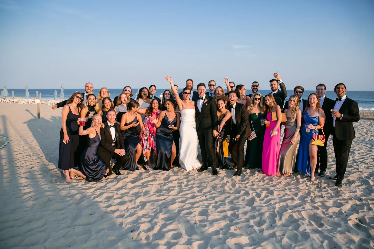 A big crowd of guests in bright dresses and tuxedos gathers around the couple on the beach