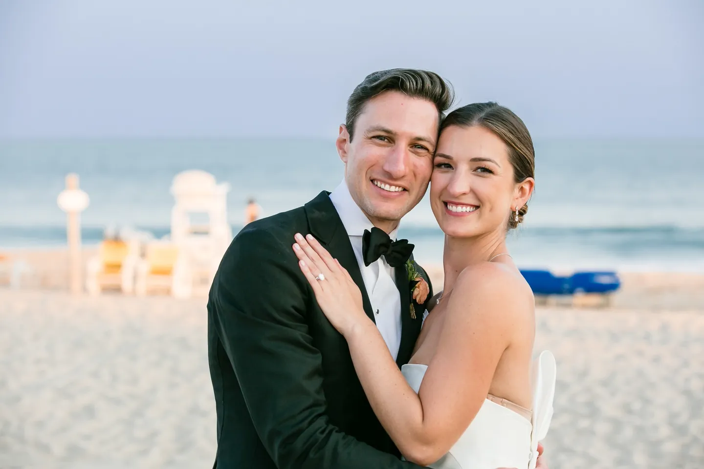 Avery and Dan press their cheeks together and smile on the beach, lifeguard stand behind