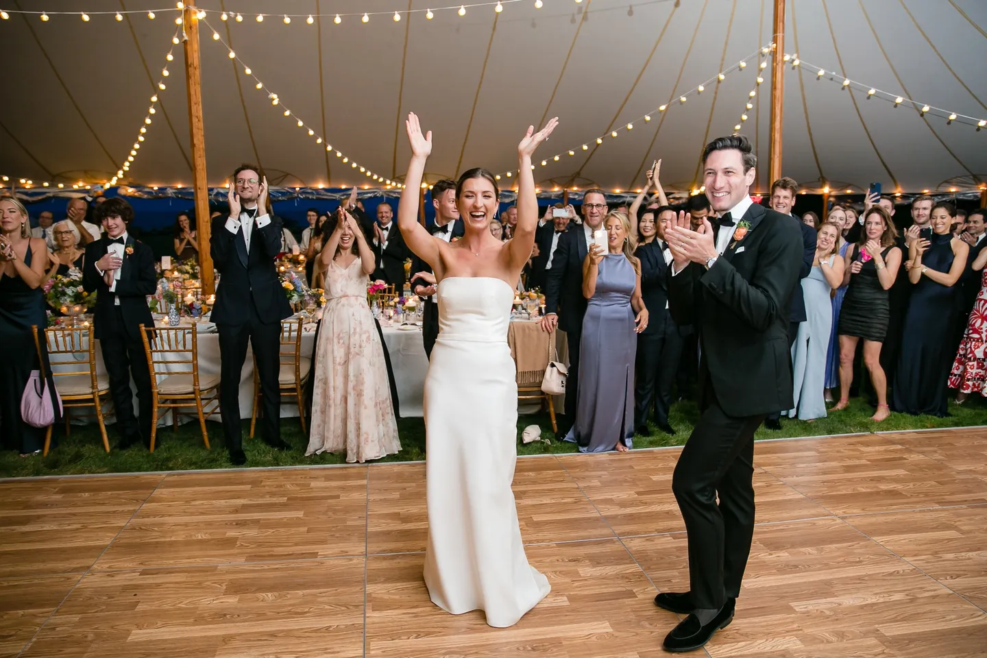 The bride throws her arms up as the couple enters the string-lit tent to a cheering crowd