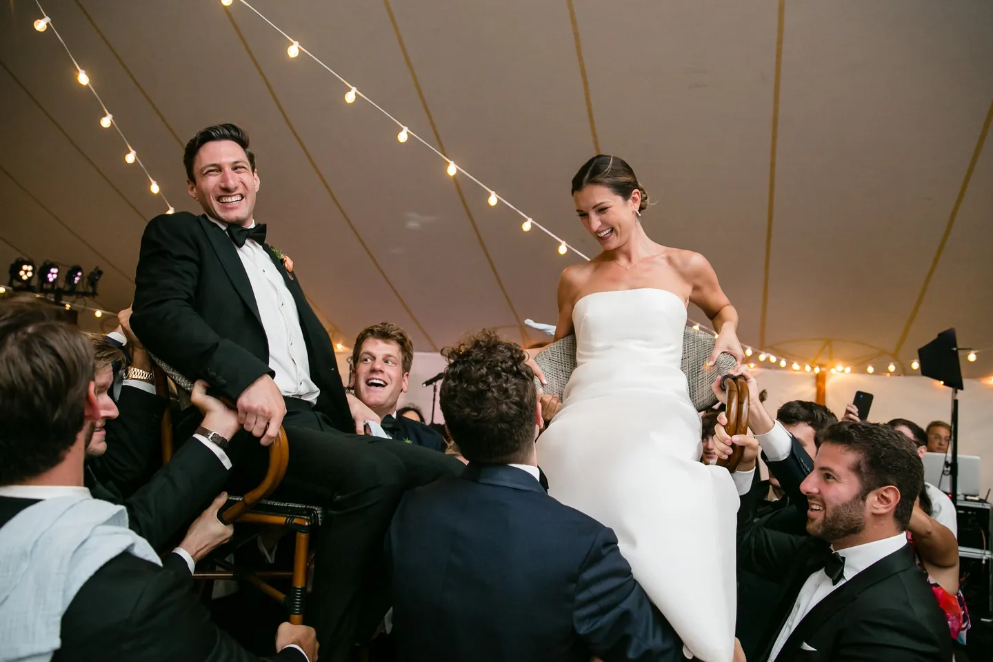 The bride is lifted high in a chair above a crowd of dancing guests under the tent