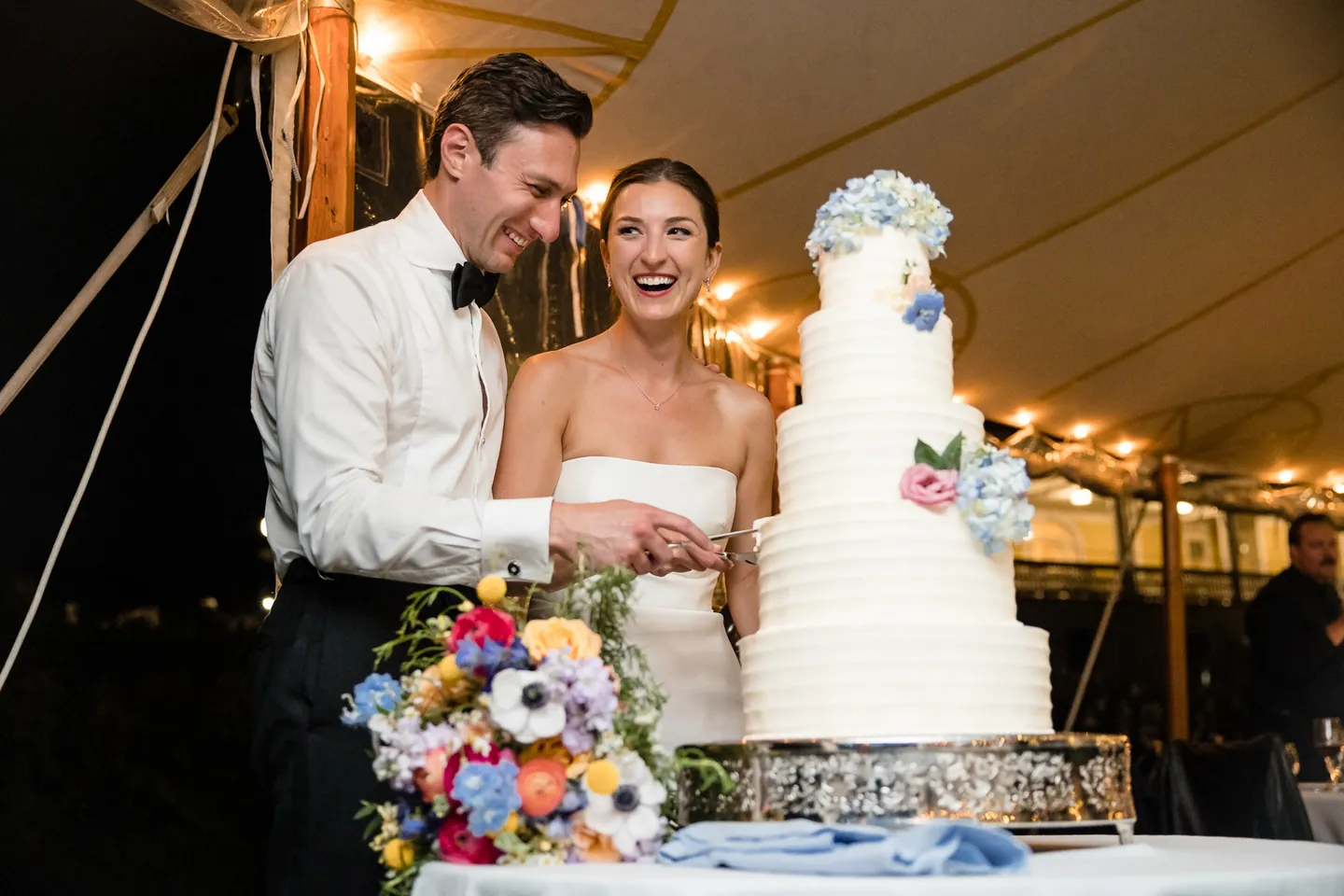 Avery and Dan laugh while cutting a tall white cake trimmed with blue flowers