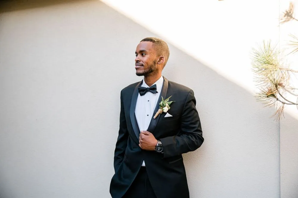The groom in a black tuxedo stands by a white wall in soft daylight