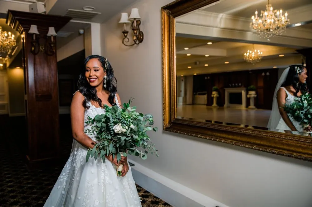 The bride holds a eucalyptus bouquet beside a gilded mirror in the hall