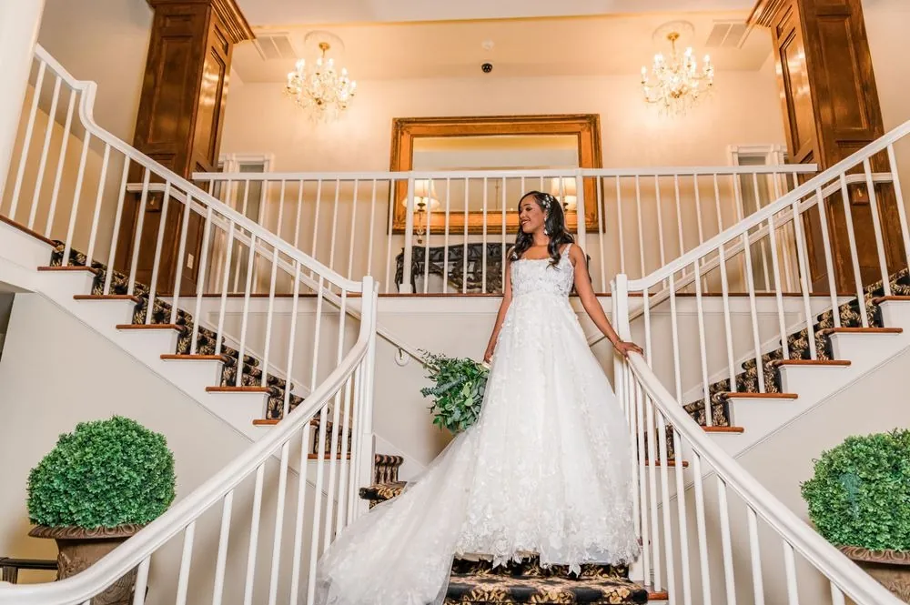 The bride stands at the foot of a white double staircase in her lace ball gown