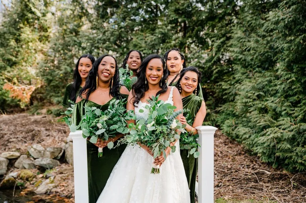Bridesmaids in olive velvet gather around the bride outdoors among evergreens