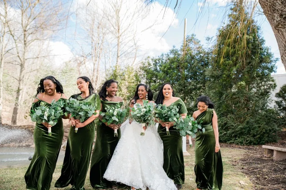 The bride laughs with bridesmaids in olive velvet under bare trees in the cold