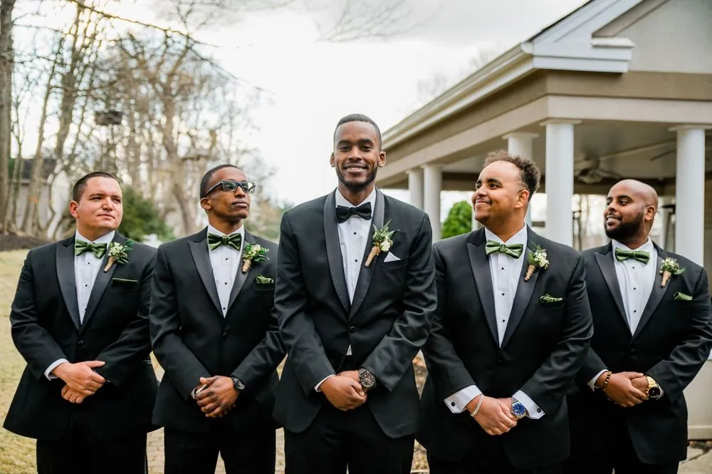 The groom and groomsmen in black tuxedos lined up in front of the white porch