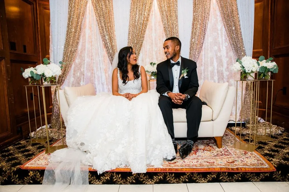 The couple sits on a cream sofa framed by gold drapes and white flowers