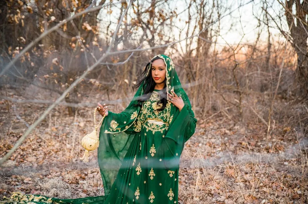 The bride in an emerald and gold embroidered gown stands in the bare winter woods