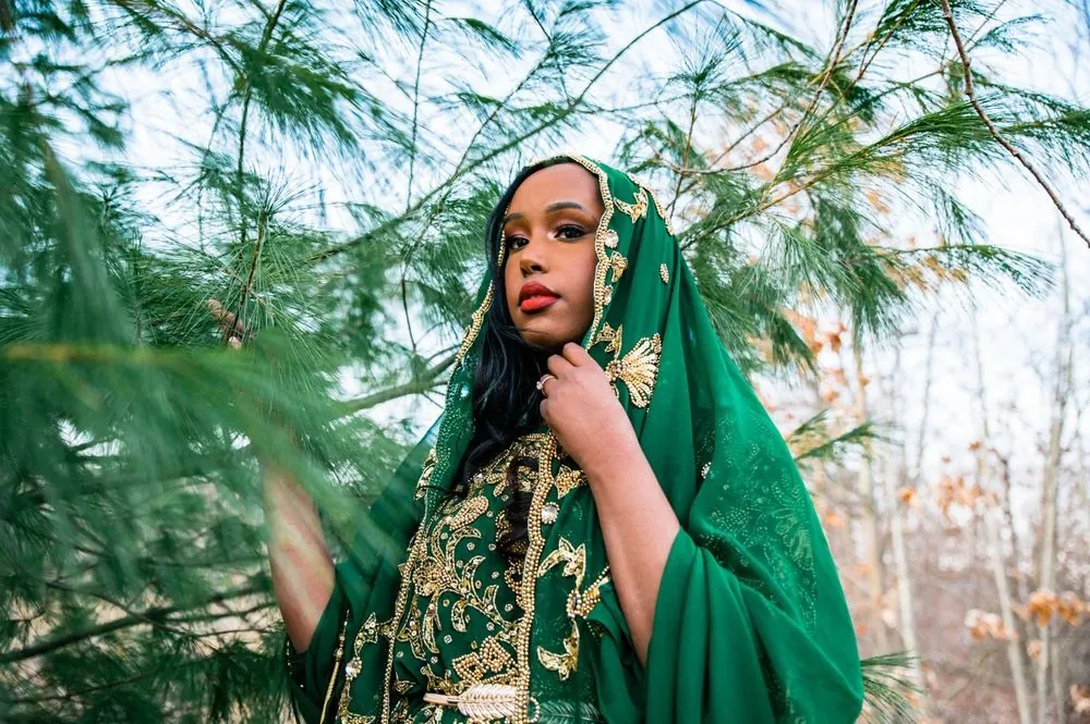 The bride in an emerald head covering with gold embroidery poses among pine branches
