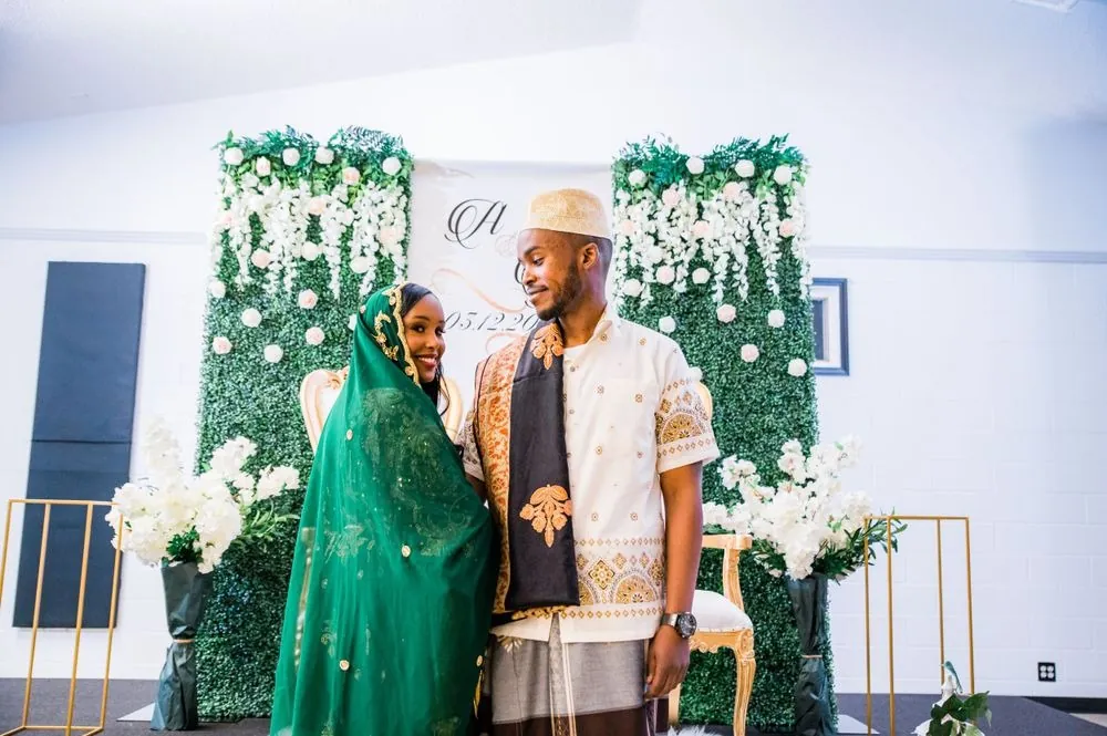 The couple in traditional attire stands before a white floral backdrop