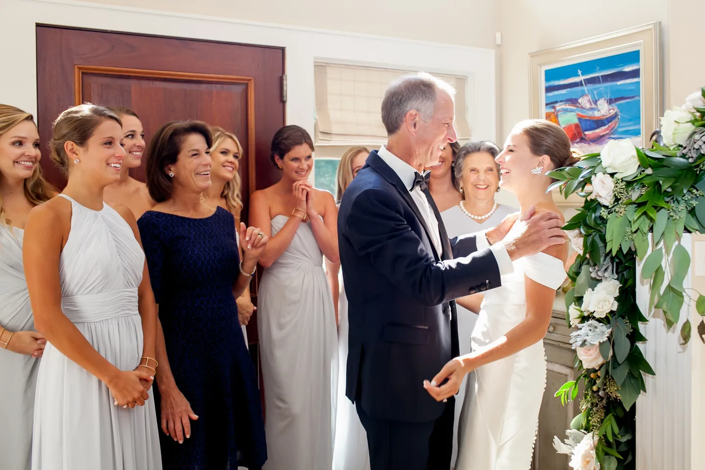 A gray-haired man in a tuxedo takes the bride's hands as bridesmaids watch in the front hall