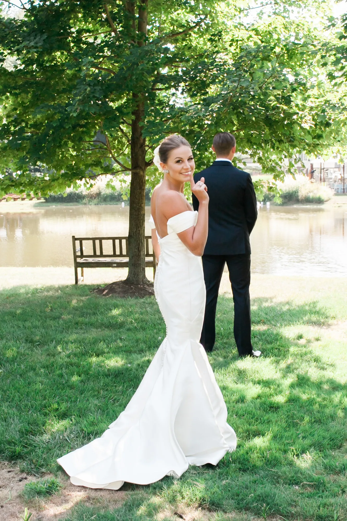 The bride walks up behind the groom under a tall shade tree on an open lawn