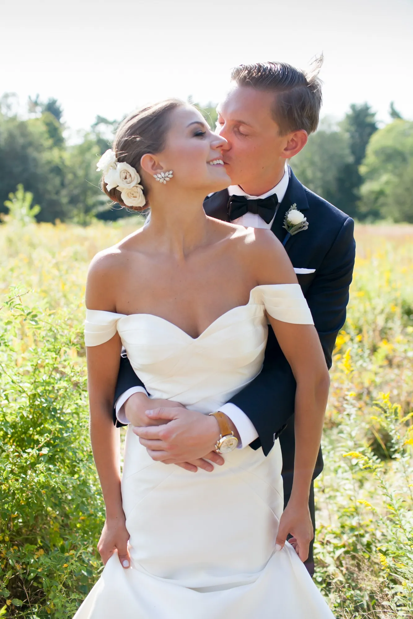 Becca and James share a kiss among green leaves, her off-shoulder gown catching the light
