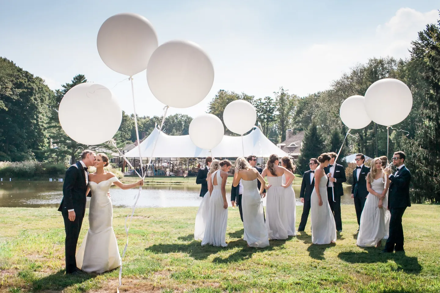 Becca and James kiss by the pond as the wedding party holds giant white balloons