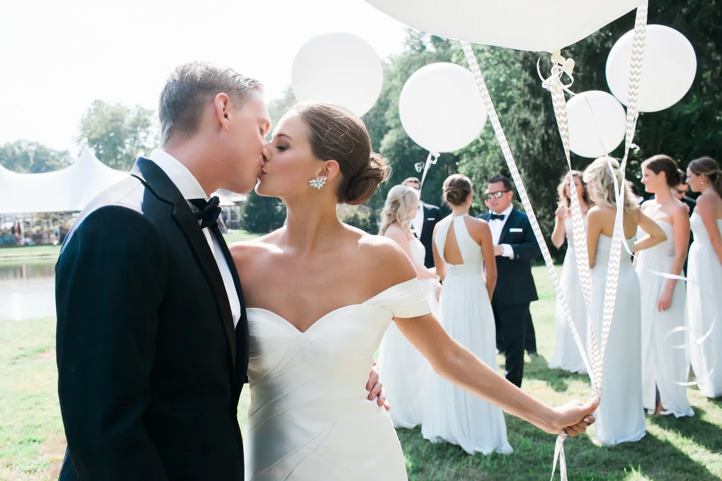 James kisses Becca as she holds balloon ribbons, bridesmaids in pale gray behind them