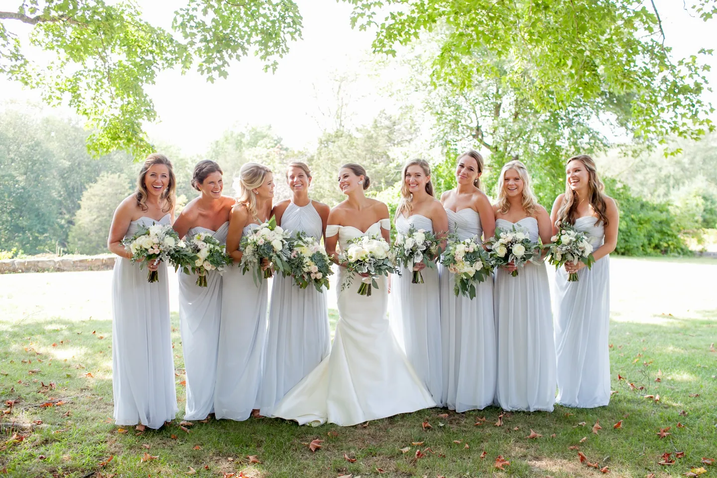 Bridesmaids in pale gray gowns stand in a row on the lawn holding bouquets