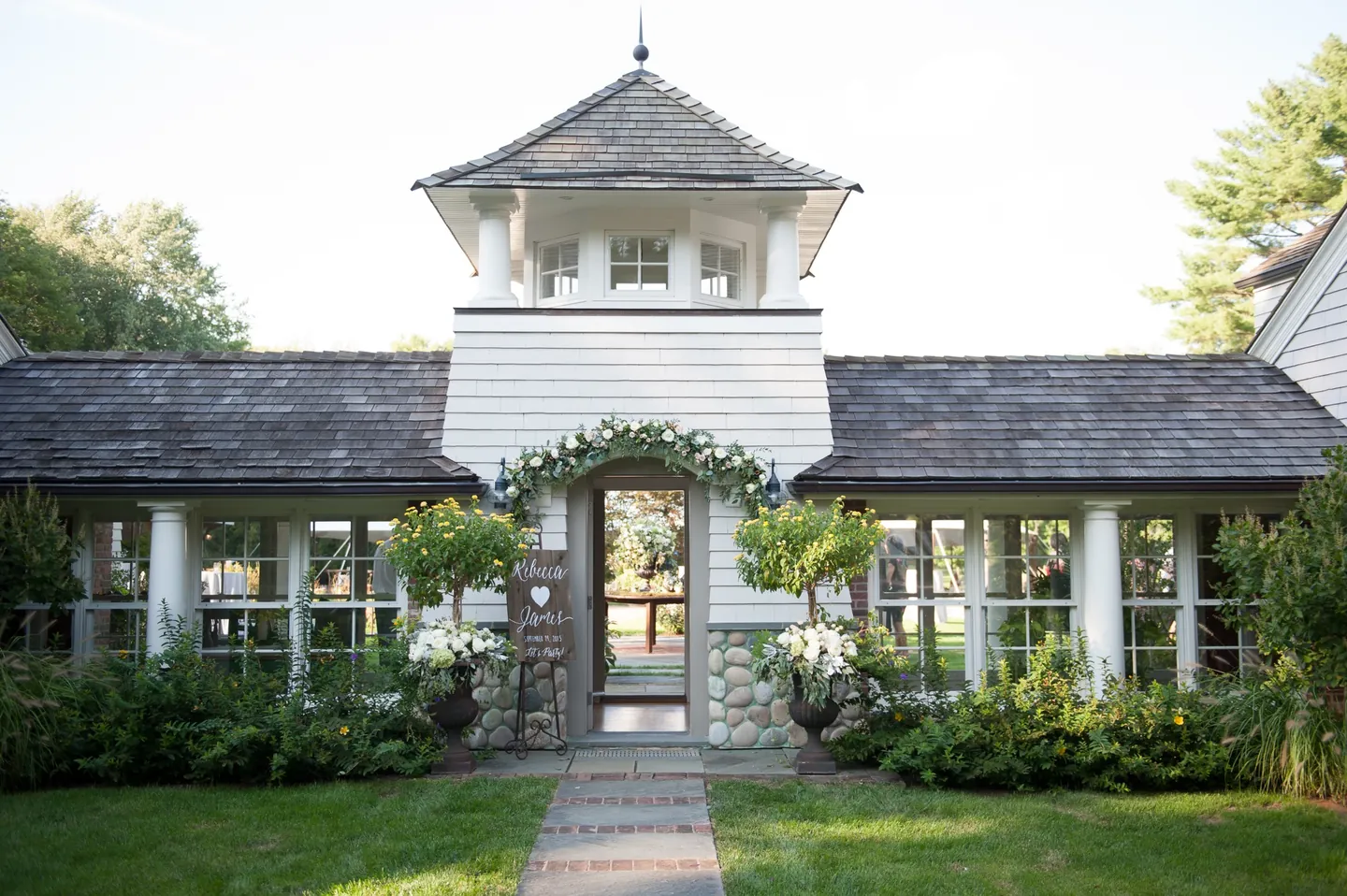 A white pool house with a cupola behind a clipped hedge and garden path
