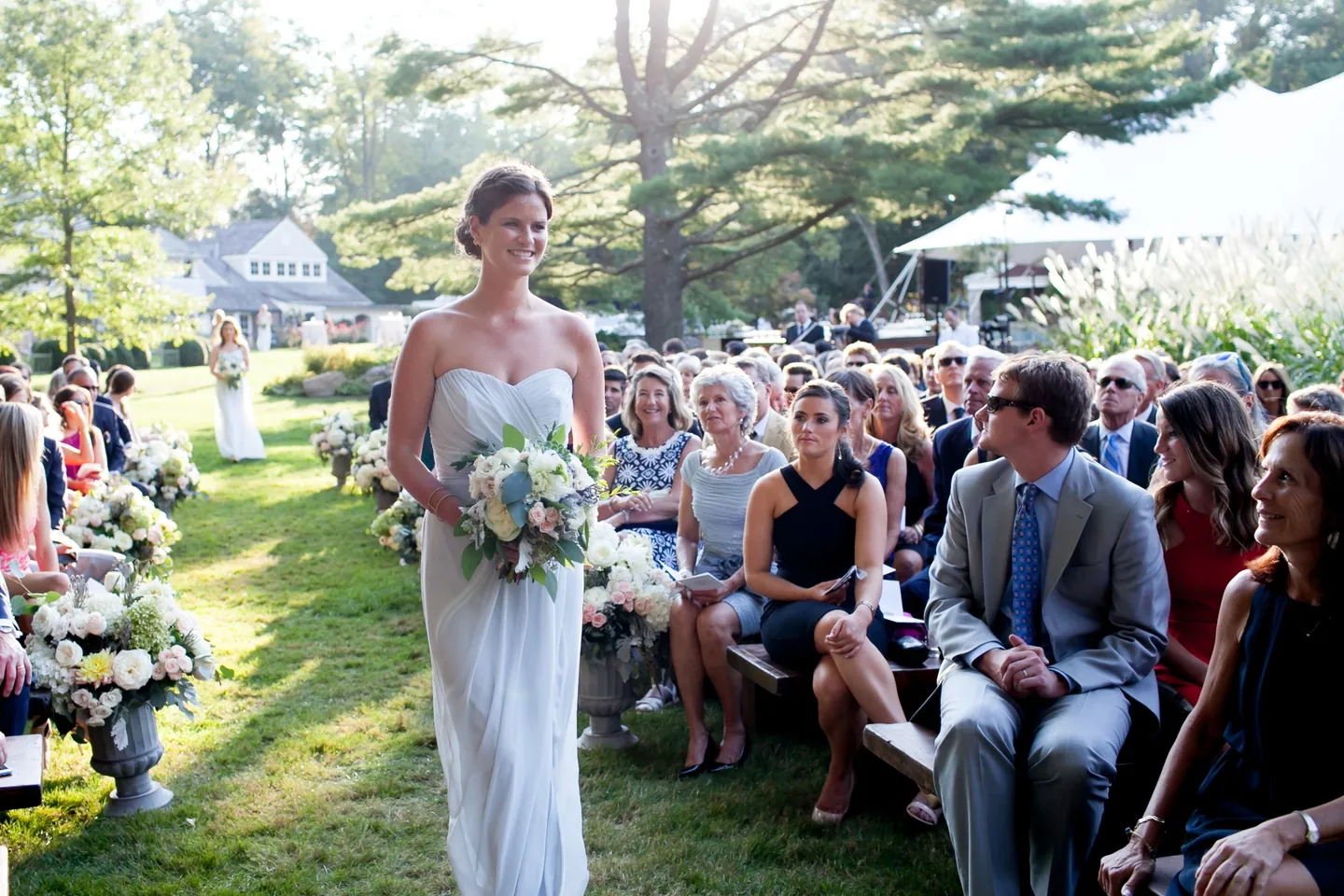 The bride walks down a grass aisle lined with white flowers as guests turn to watch