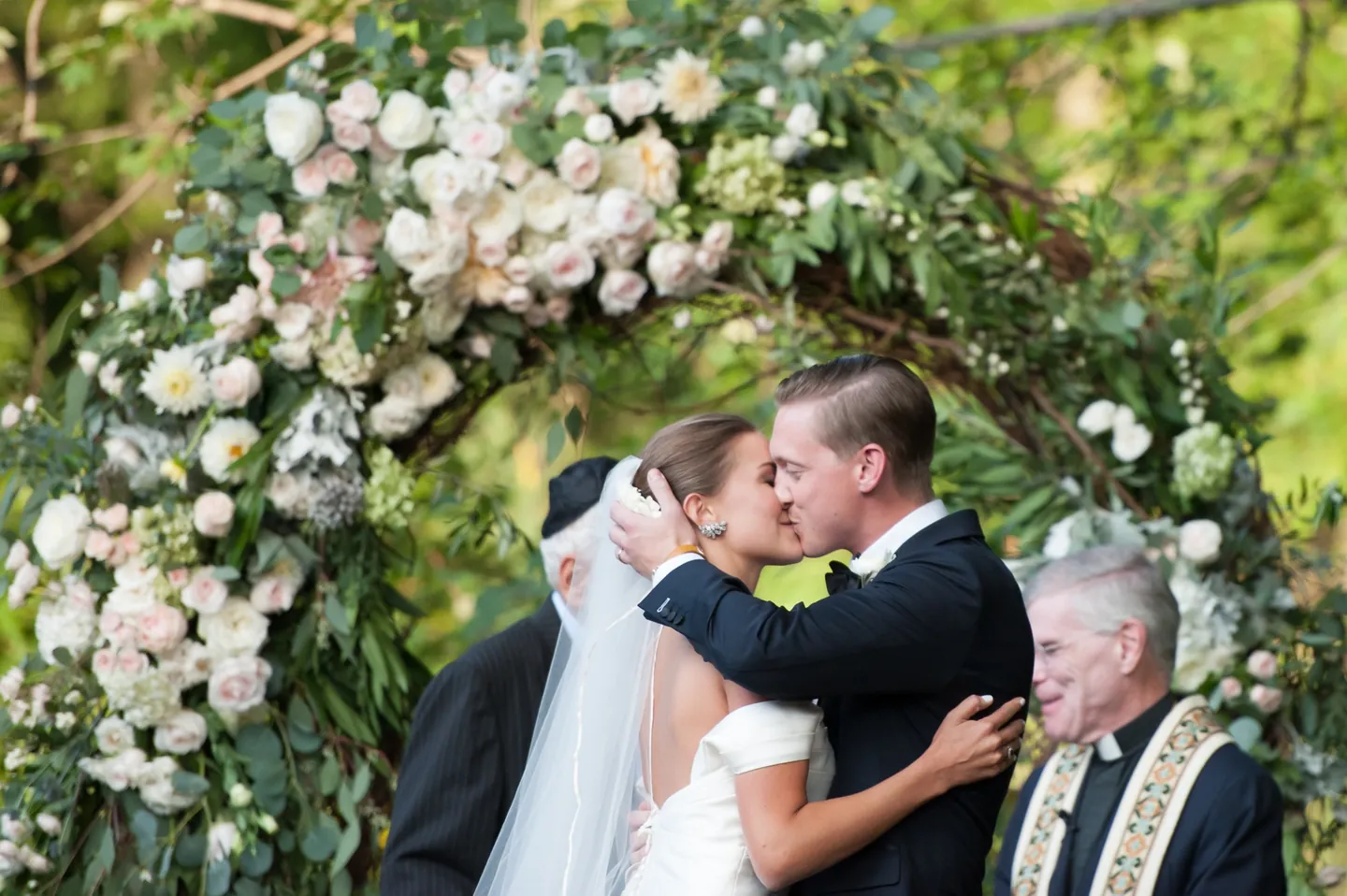 Becca and James kiss under an arch of blush roses as an officiant smiles
