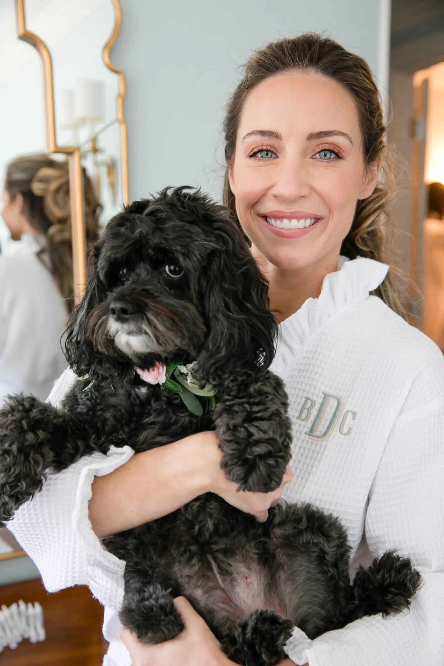 The bride in a white robe cuddles a small black curly-haired dog and smiles