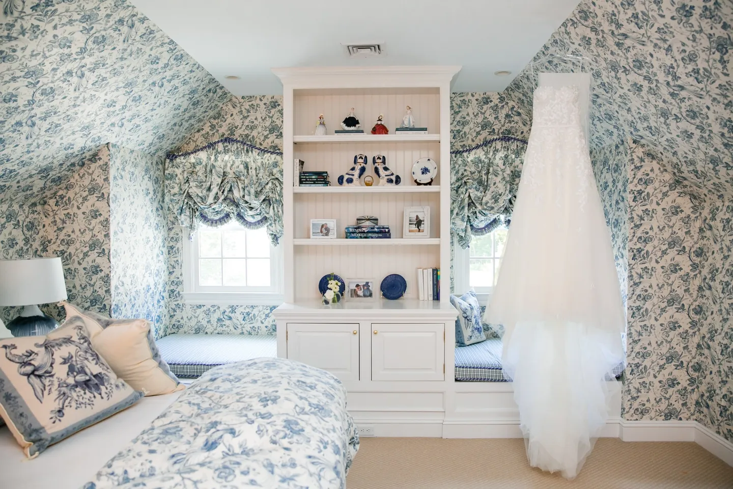 A lace gown hangs in a bedroom papered in blue toile with a window seat