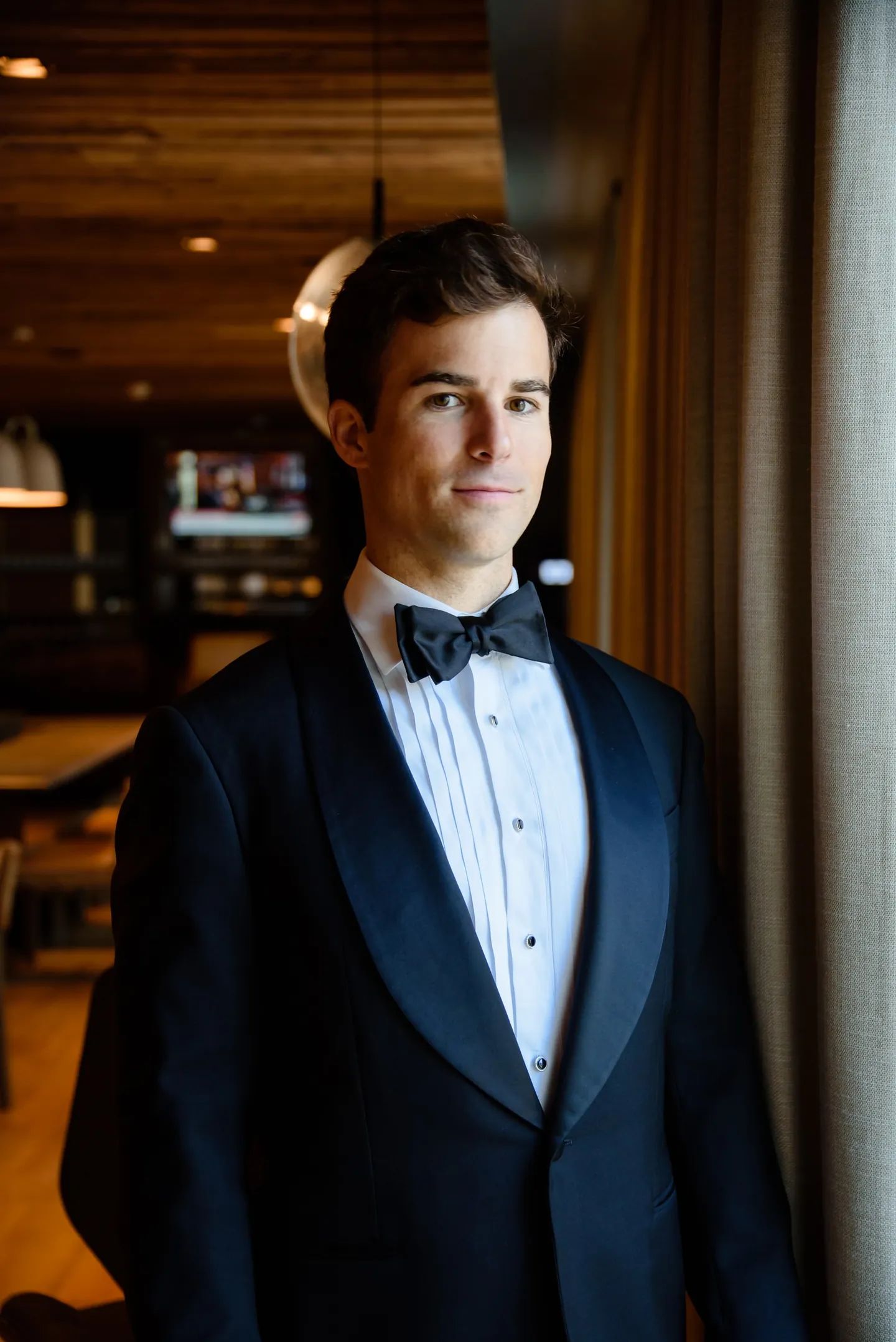 The groom stands in a midnight tux with a bow tie in a dark wood-paneled room