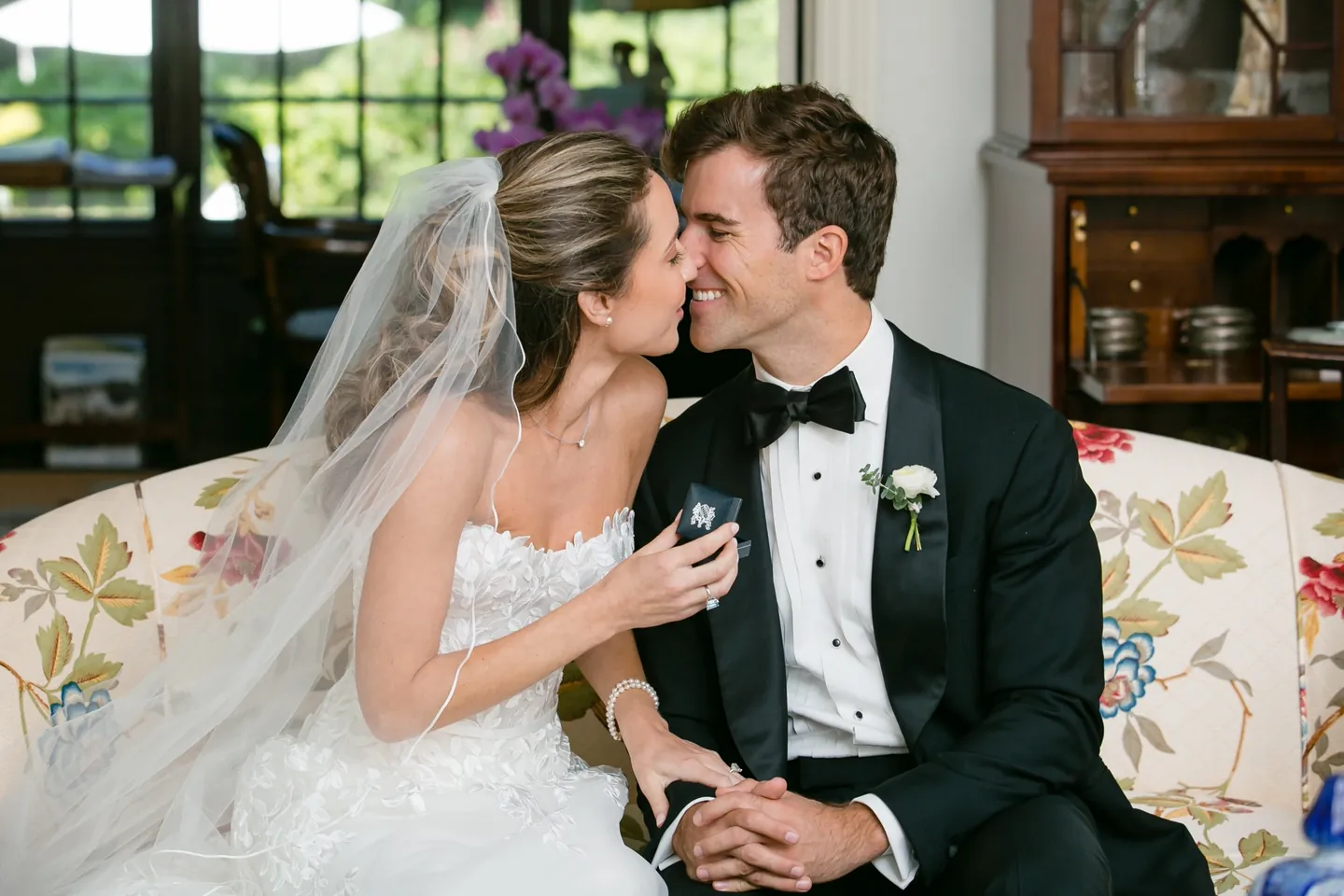 Britt and Andrew lean close together on a floral sofa, her veil draped around them