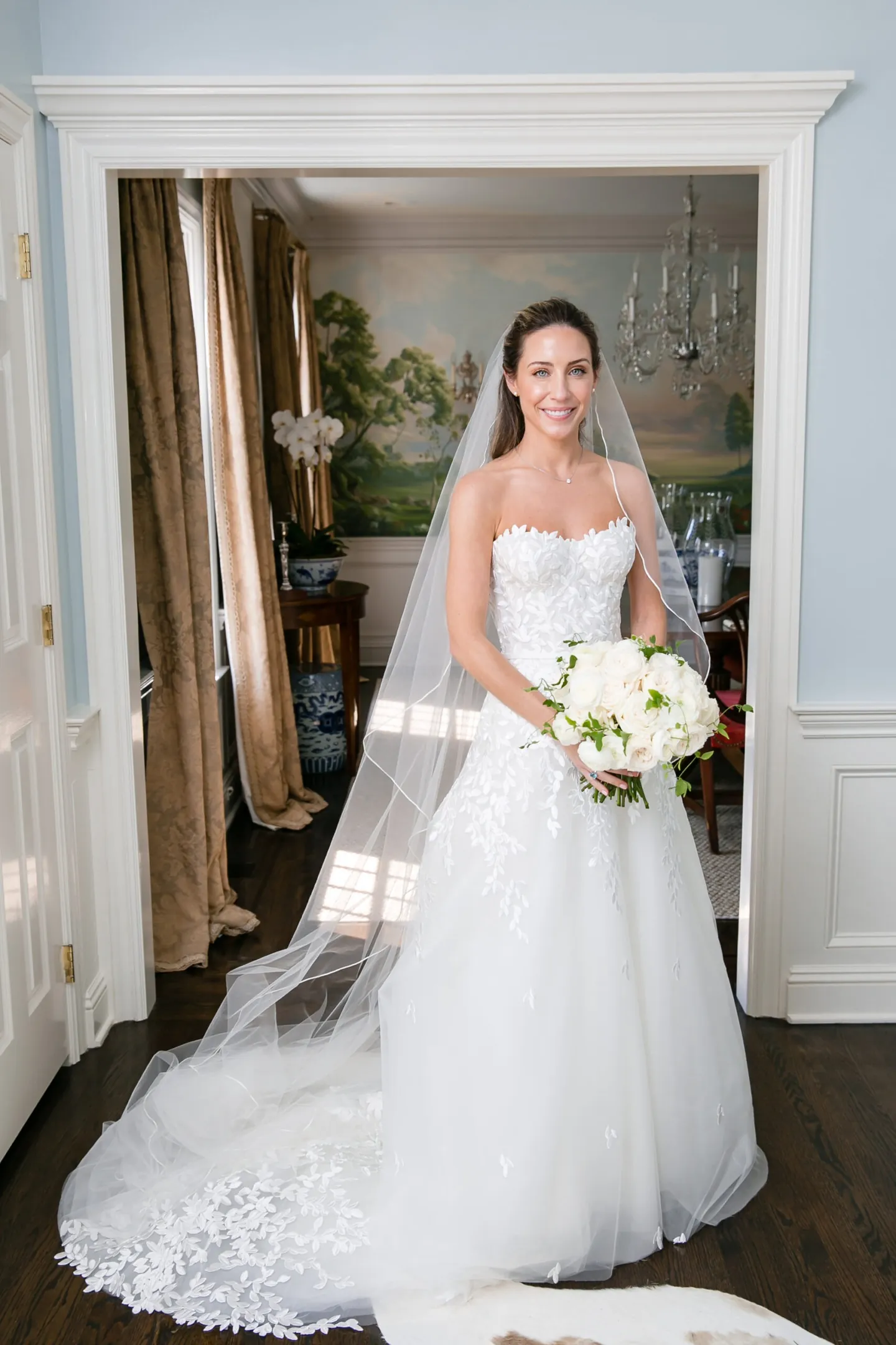 The bride stands in a white doorway holding a white bouquet, veil falling behind her