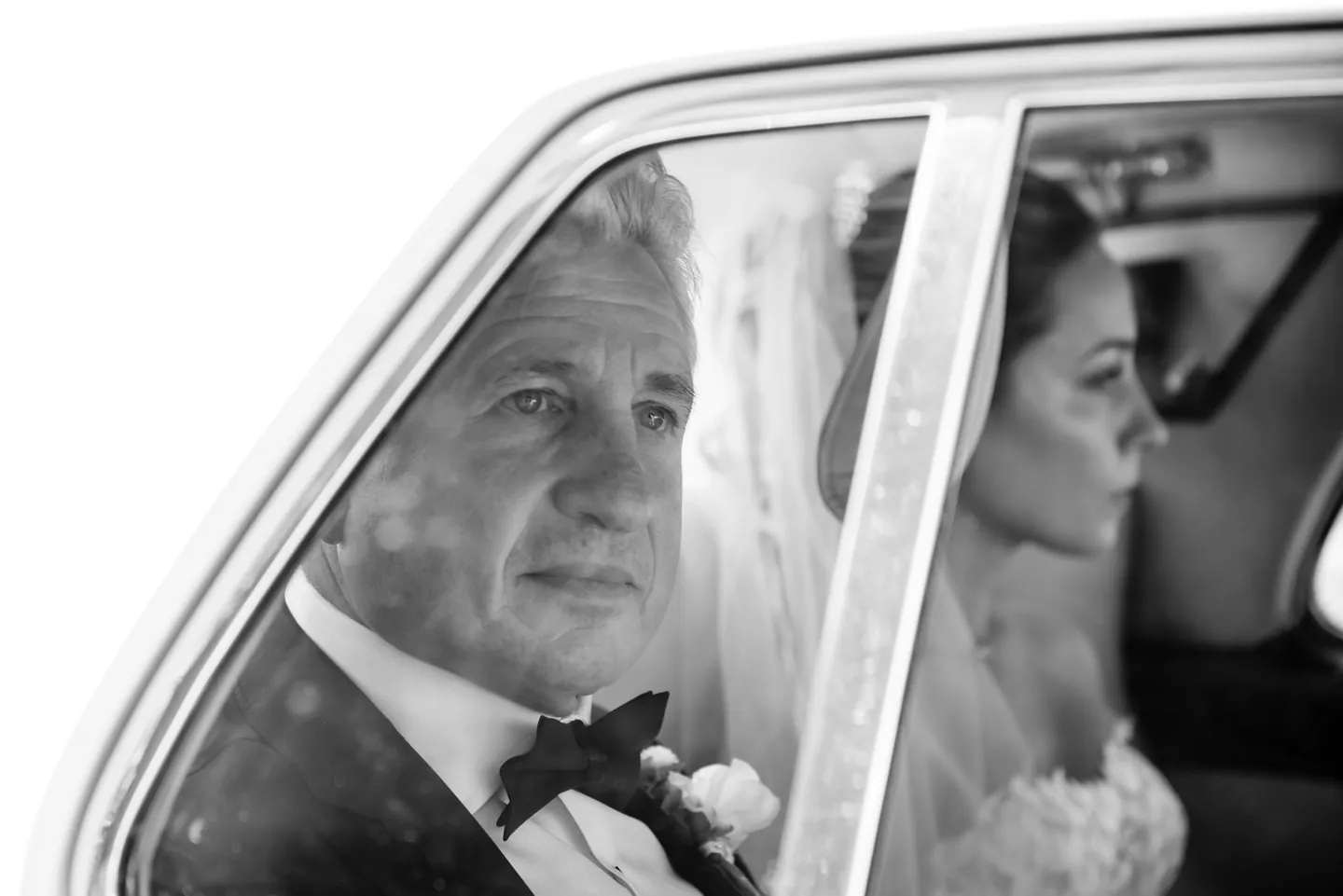 Black and white frame through a car window of an older man in a tux beside the bride