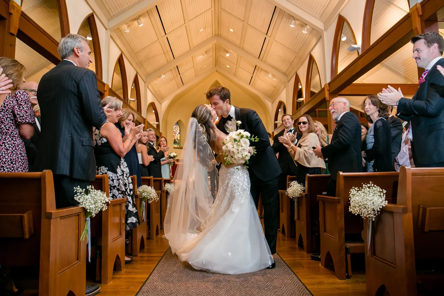 Britt and Andrew kiss in the chapel aisle under a warm wood ceiling as guests clap